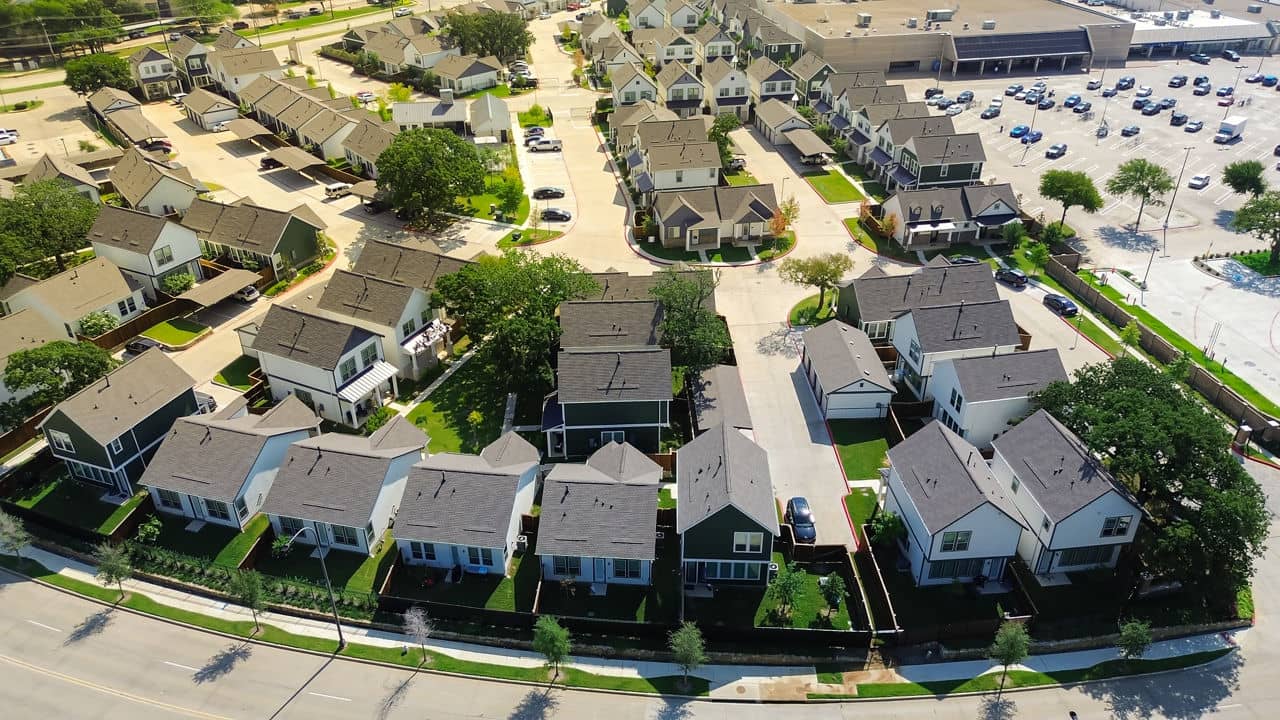 Aerial view of homes in Northeast Dallas, Texas, featuring green lawns and trees, with a shopping area in the background.
