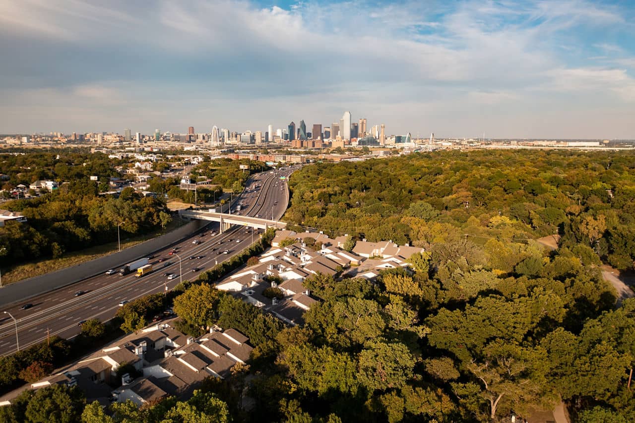 Aerial view of Oak Cliff, Texas, with a highway and the Dallas skyline in the background surrounded by trees.