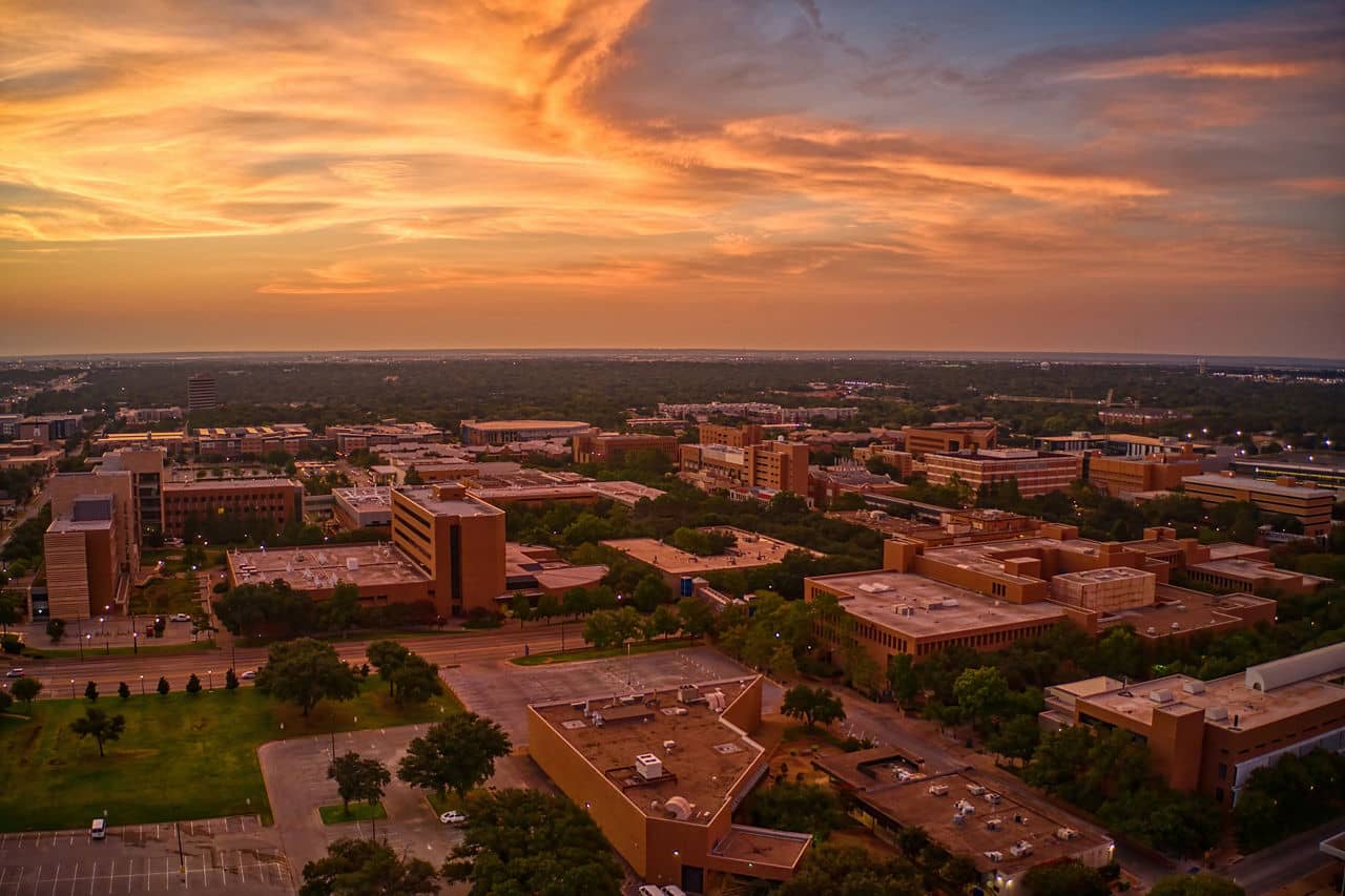 Aerial view of Arlington, Texas at sunset, showcasing buildings and green spaces under a colorful sky.