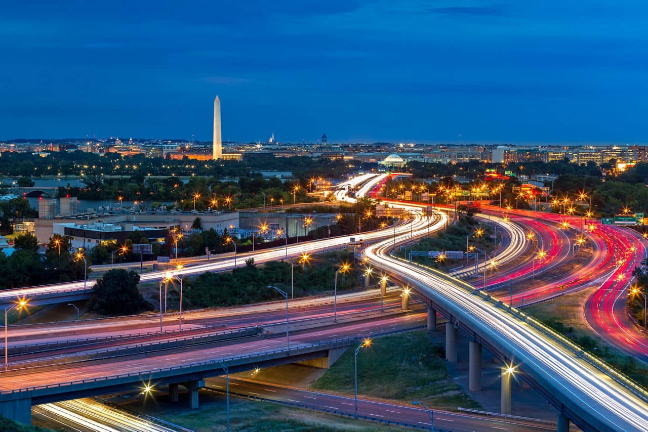 Washington D.C. cityscape at dusk with rush hour traffic trails on I-395 highway. Washington Monument, illuminated, dominates the skyline.