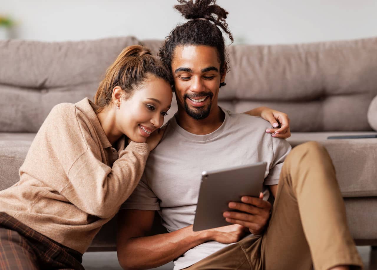 Young lovely african american family couple sitting on floor with digital tablet with smile on face while spending great time together at home, happy husband and wife watching funny video on computer
